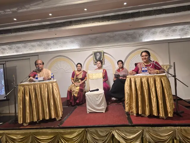 Performers seated on a stage with gold-draped tables beneath a portrait of Tagore