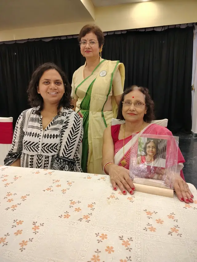 A host stands behind two seated women holding a framed portrait